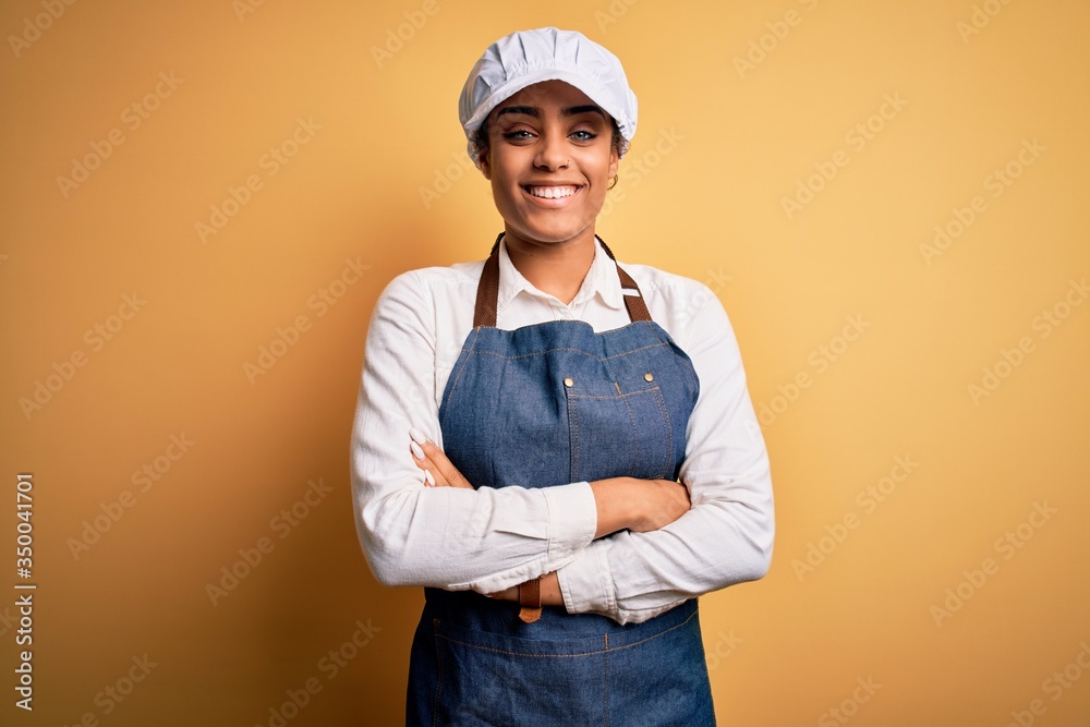 Young african american afro baker woman wearing apron and cap over yellow background happy face smiling with crossed arms looking at the camera. Positive person. Young african american afro baker woman wearing apron and cap over yellow background happy face smiling with crossed arms looking at the camera. Positive person.