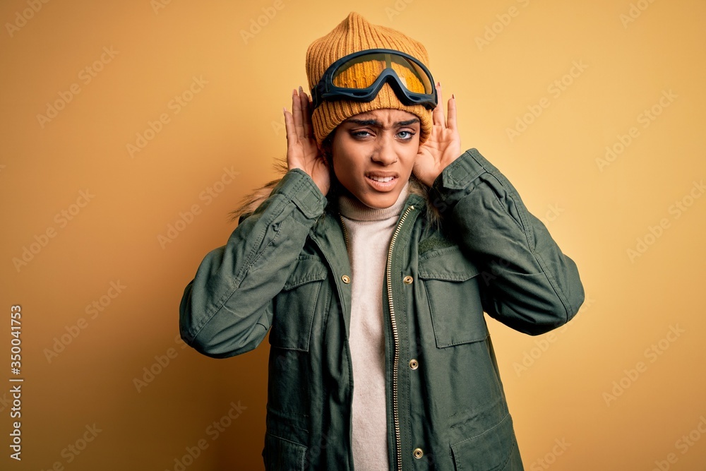 Young african american afro skier girl wearing snow sportswear and ski goggles Trying to hear both hands on ear gesture, curious for gossip. Hearing problem, deaf Young african american afro skier girl wearing snow sportswear and ski goggles Trying to hear both hands on ear gesture, curious for gossip. Hearing problem, deaf