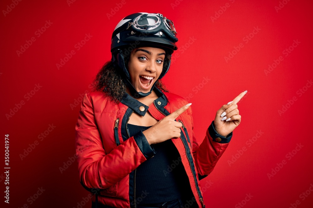 Young african american motorcyclist girl wearing moto helmet and glasses over red background smiling and looking at the camera pointing with two hands and fingers to the side. Young african american motorcyclist girl wearing moto helmet and glasses over red background smiling and looking at the camera pointing with two hands and fingers to the side.