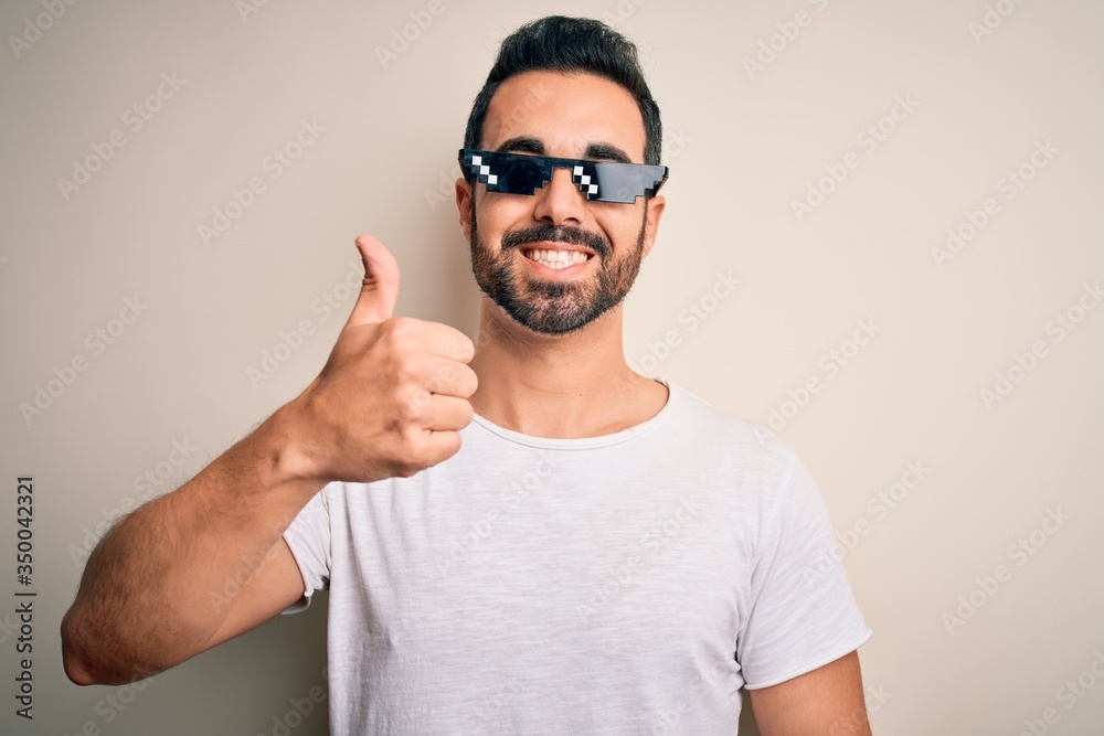 Young handsome man with beard wearing funny thug life sunglasses over white background doing happy thumbs up gesture with hand. Approving expression looking at the camera showing success. Young handsome man with beard wearing funny thug life sunglasses over white background doing happy thumbs up gesture with hand. Approving expression looking at the camera showing success.