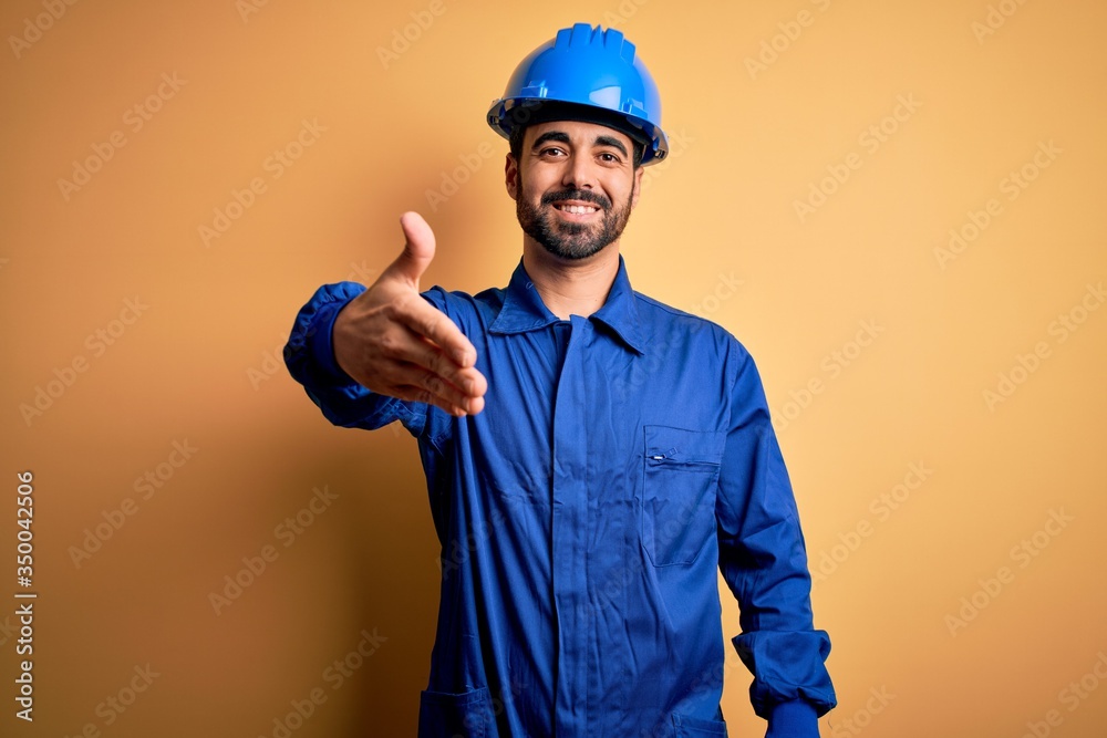 Mechanic man with beard wearing blue uniform and safety helmet over yellow background smiling friendly offering handshake as greeting and welcoming. Successful business.