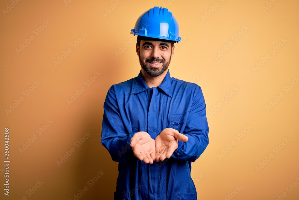 Mechanic man with beard wearing blue uniform and safety helmet over yellow background Smiling with hands palms together receiving or giving gesture. Hold and protection