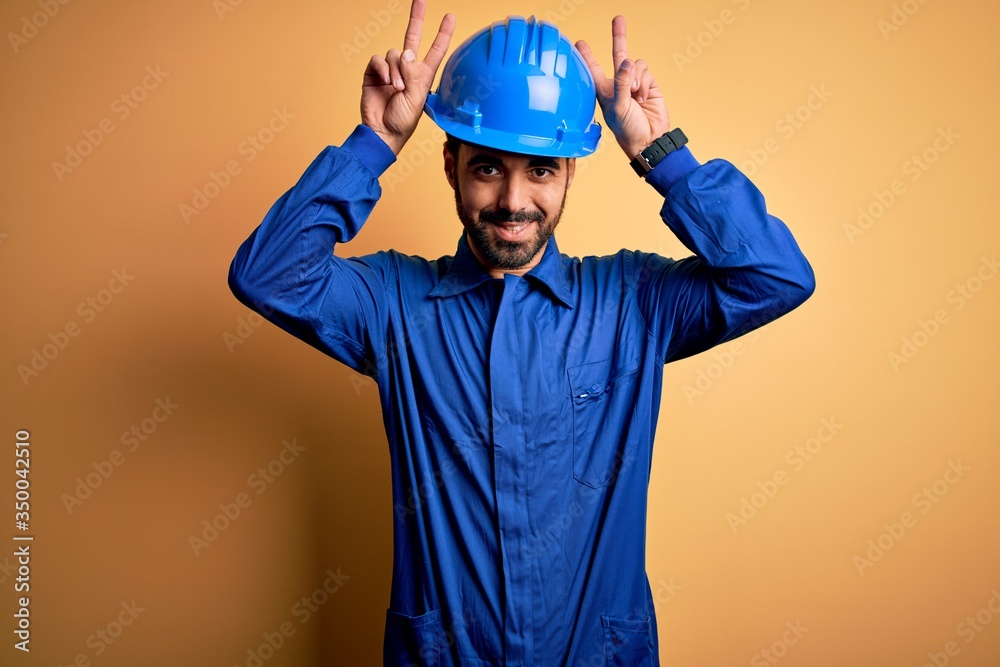 Mechanic man with beard wearing blue uniform and safety helmet over yellow background Posing funny and crazy with fingers on head as bunny ears, smiling cheerful