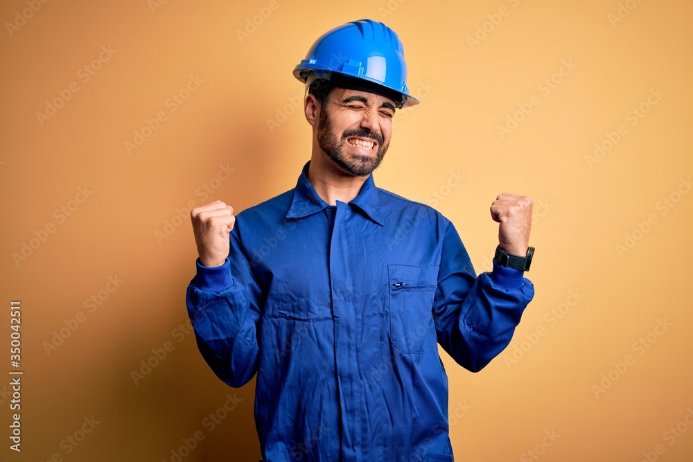 Mechanic man with beard wearing blue uniform and safety helmet over yellow background very happy and excited doing winner gesture with arms raised, smiling and screaming for success. Celebration