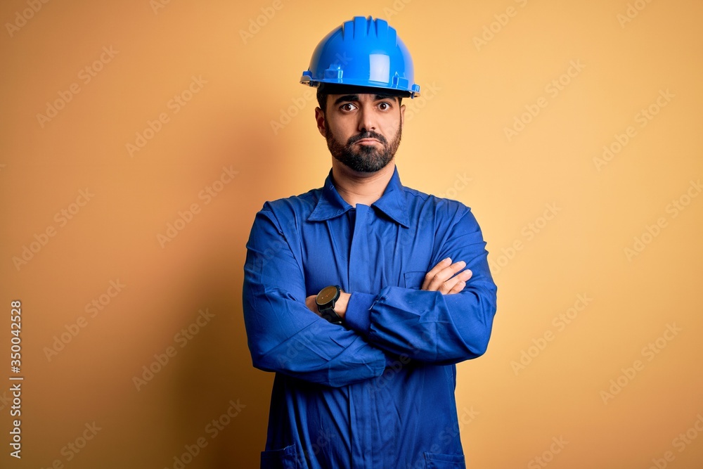 Mechanic man with beard wearing blue uniform and safety helmet over yellow background skeptic and nervous, disapproving expression on face with crossed arms. Negative person.