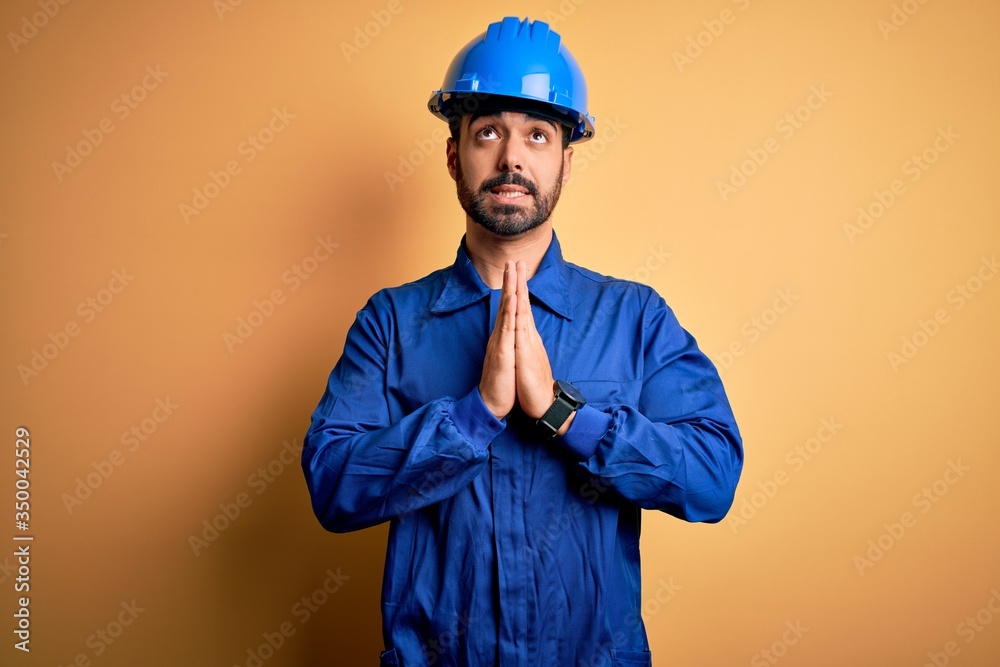 Mechanic man with beard wearing blue uniform and safety helmet over yellow background begging and praying with hands together with hope expression on face very emotional and worried. Begging.