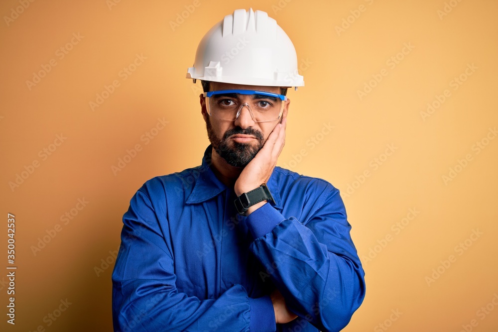 Mechanic man with beard wearing blue uniform and safety glasses over yellow background thinking looking tired and bored with depression problems with crossed arms.