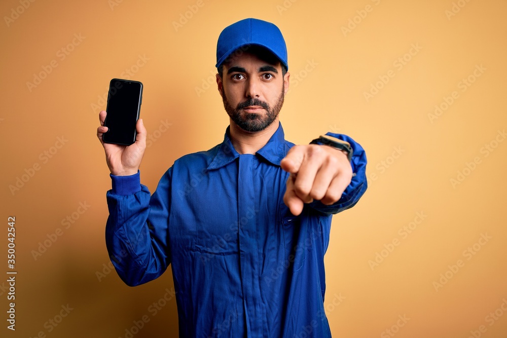 Mechanic man with beard wearing blue uniform and cap holding smartphone showing screen pointing with finger to the camera and to you, hand sign, positive and confident gesture from the front