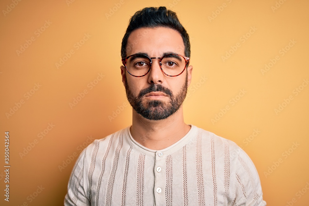 Young handsome man with beard wearing casual t-shirt and glasses over yellow background with serious expression on face. Simple and natural looking at the camera. Young handsome man with beard wearing casual t-shirt and glasses over yellow background with serious expression on face. Simple and natural looking at the camera.