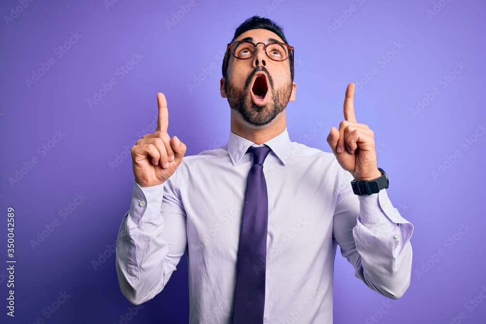 Handsome businessman with beard wearing casual tie and glasses over purple background amazed and surprised looking up and pointing with fingers and raised arms. Handsome businessman with beard wearing casual tie and glasses over purple background amazed and surprised looking up and pointing with fingers and raised arms.