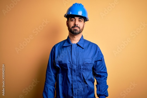 Mechanic man with beard wearing blue uniform and safety helmet over yellow background looking sleepy and tired, exhausted for fatigue and hangover, lazy eyes in the morning.
