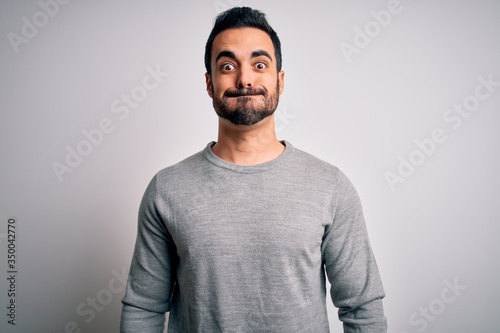 Young handsome man with beard wearing casual sweater standing over white background puffing cheeks with funny face. Mouth inflated with air, crazy expression.