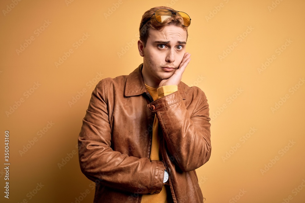 Young handsome redhead man wearing casual leather jacket over isolated yellow background thinking looking tired and bored with depression problems with crossed arms. Young handsome redhead man wearing casual leather jacket over isolated yellow background thinking looking tired and bored with depression problems with crossed arms.
