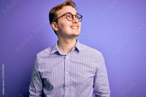 Young handsome redhead man wearing casual shirt and glasses over purple background looking away to side with smile on face, natural expression. Laughing confident. © Krakenimages.com