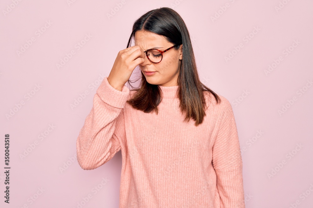 Young beautiful brunette woman wearing casual sweater and glasses over pink background tired rubbing nose and eyes feeling fatigue and headache. Stress and frustration concept. Young beautiful brunette woman wearing casual sweater and glasses over pink background tired rubbing nose and eyes feeling fatigue and headache. Stress and frustration concept.