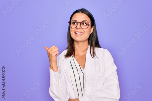 Young beautiful brunette psychologist woman wearing coat and glasses over purple background smiling with happy face looking and pointing to the side with thumb up.
