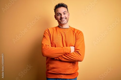 Young handsome man wearing orange casual sweater standing over isolated yellow background happy face smiling with crossed arms looking at the camera. Positive person.