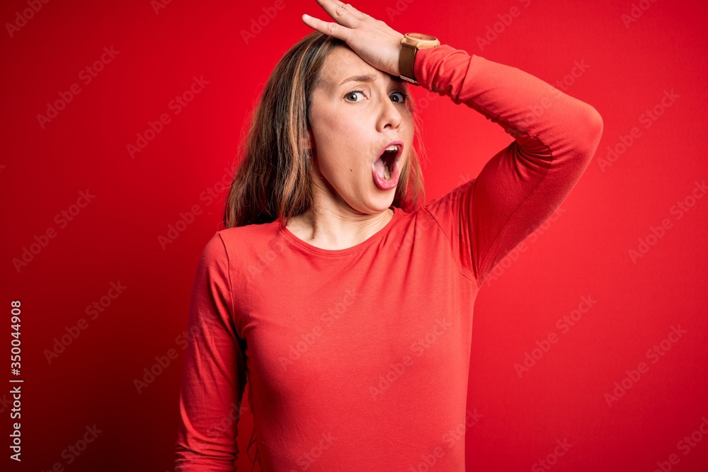 Young beautiful blonde woman wearing casual t-shirt standing over isolated red background surprised with hand on head for mistake, remember error. Forgot, bad memory concept. Young beautiful blonde woman wearing casual t-shirt standing over isolated red background surprised with hand on head for mistake, remember error. Forgot, bad memory concept.