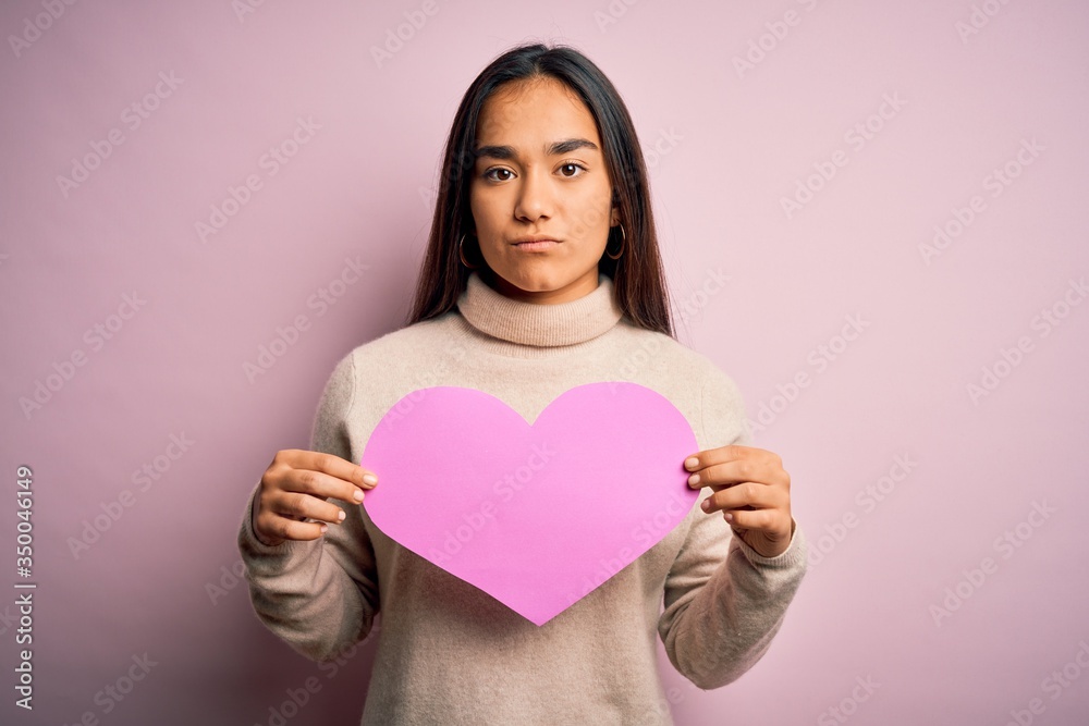 Young beautiful asian woman holding pink heart standing over isolated background with a confident expression on smart face thinking serious Young beautiful asian woman holding pink heart standing over isolated background with a confident expression on smart face thinking serious