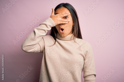 Young beautiful asian woman wearing casual turtleneck sweater over pink background peeking in shock covering face and eyes with hand, looking through fingers with embarrassed expression. © Krakenimages.com