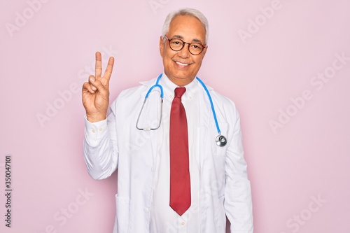 Middle age senior grey-haired doctor man wearing stethoscope and professional medical coat smiling looking to the camera showing fingers doing victory sign. Number two. © Krakenimages.com