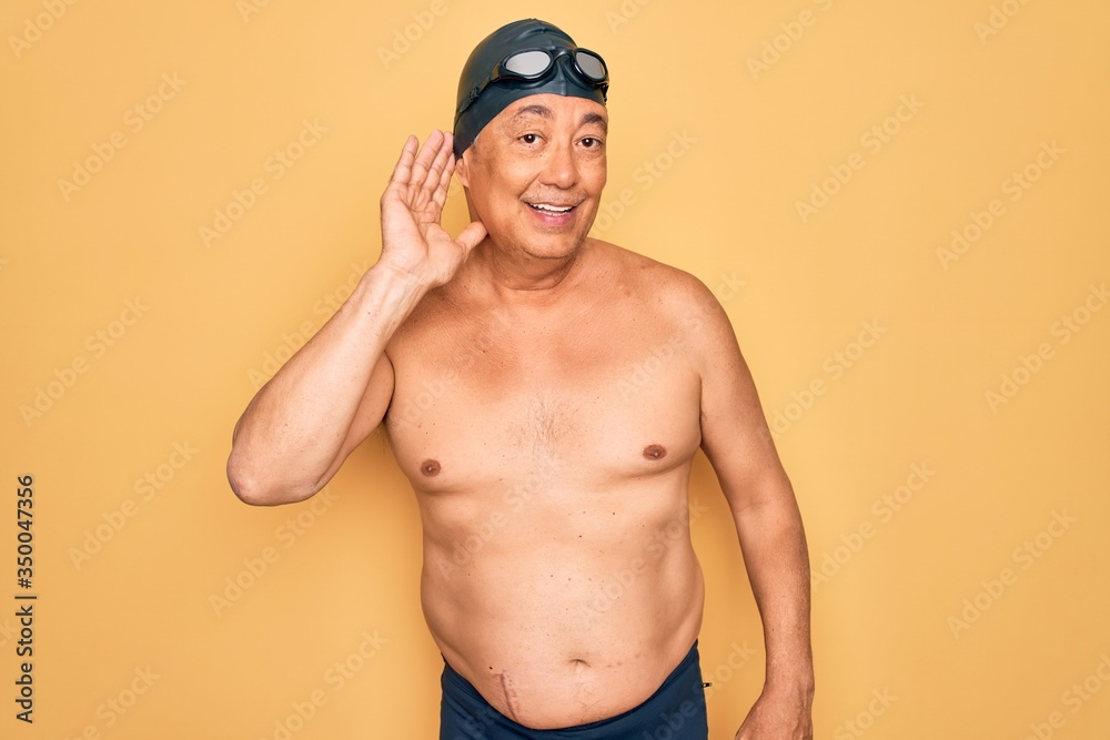 Middle age senior grey-haired swimmer man wearing swimsuit, cap and goggles smiling with hand over ear listening an hearing to rumor or gossip. Deafness concept.