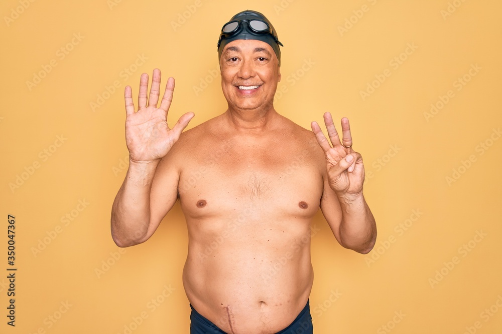 Middle age senior grey-haired swimmer man wearing swimsuit, cap and goggles showing and pointing up with fingers number eight while smiling confident and happy.
