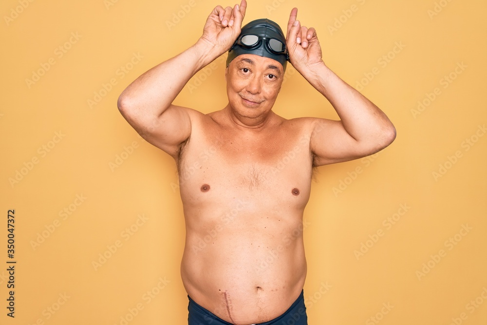 Middle age senior grey-haired swimmer man wearing swimsuit, cap and goggles doing funny gesture with finger over head as bull horns