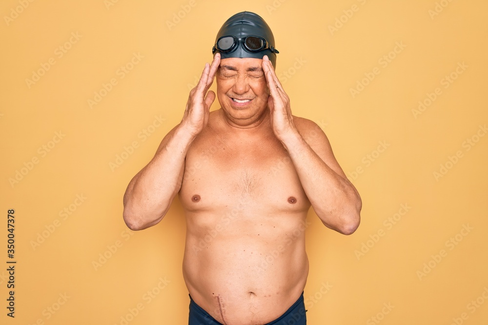 Middle age senior grey-haired swimmer man wearing swimsuit, cap and goggles with hand on headache because stress. Suffering migraine.