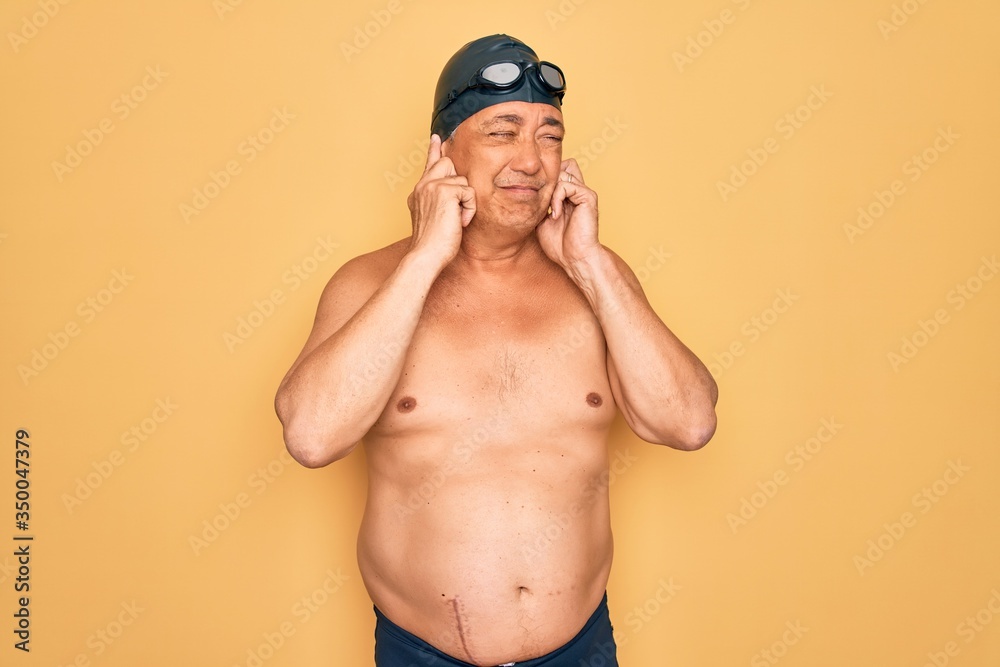 Middle age senior grey-haired swimmer man wearing swimsuit, cap and goggles covering ears with fingers with annoyed expression for the noise of loud music. Deaf concept.
