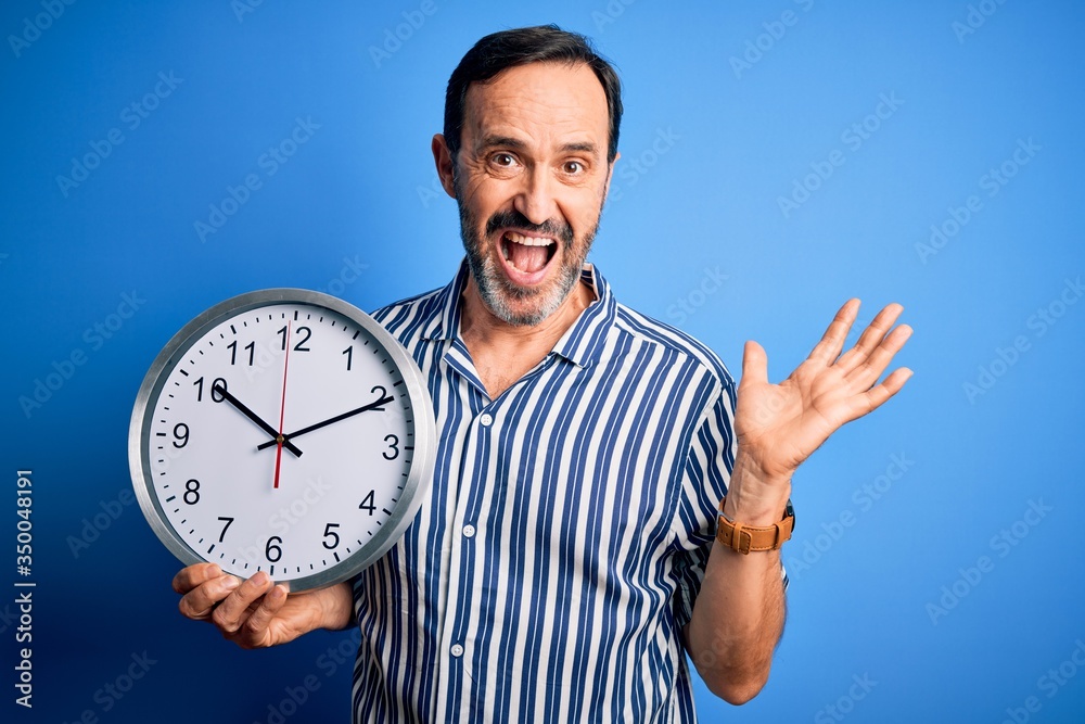 Middle age hoary man holding big clock standing over isolated blue background very happy and excited, winner expression celebrating victory screaming with big smile and raised hands Middle age hoary man holding big clock standing over isolated blue background very happy and excited, winner expression celebrating victory screaming with big smile and raised hands