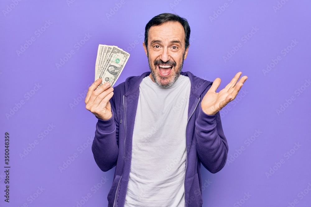 Middle age handsome man holding bunch of dollars banknotes over isolated purple background celebrating achievement with happy smile and winner expression with raised hand Middle age handsome man holding bunch of dollars banknotes over isolated purple background celebrating achievement with happy smile and winner expression with raised hand