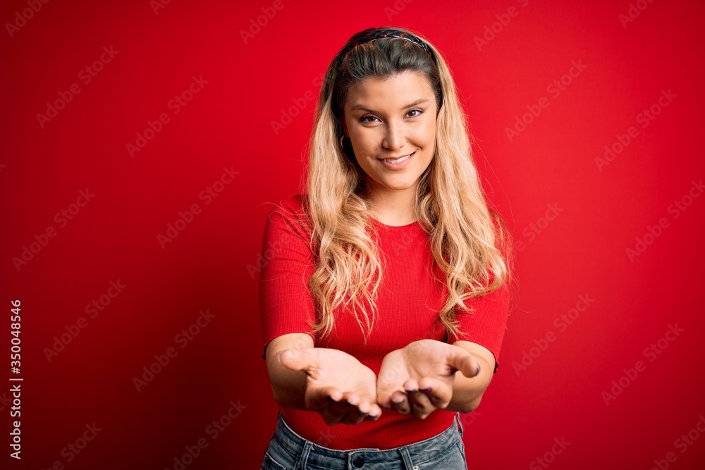 Young beautiful blonde woman wearing casual t-shirt standing over isolated red background Smiling with hands palms together receiving or giving gesture. Hold and protection Young beautiful blonde woman wearing casual t-shirt standing over isolated red background Smiling with hands palms together receiving or giving gesture. Hold and protection