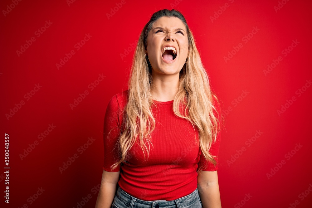 Young beautiful blonde woman wearing casual t-shirt standing over isolated red background angry and mad screaming frustrated and furious, shouting with anger. Rage and aggressive concept. Young beautiful blonde woman wearing casual t-shirt standing over isolated red background angry and mad screaming frustrated and furious, shouting with anger. Rage and aggressive concept.