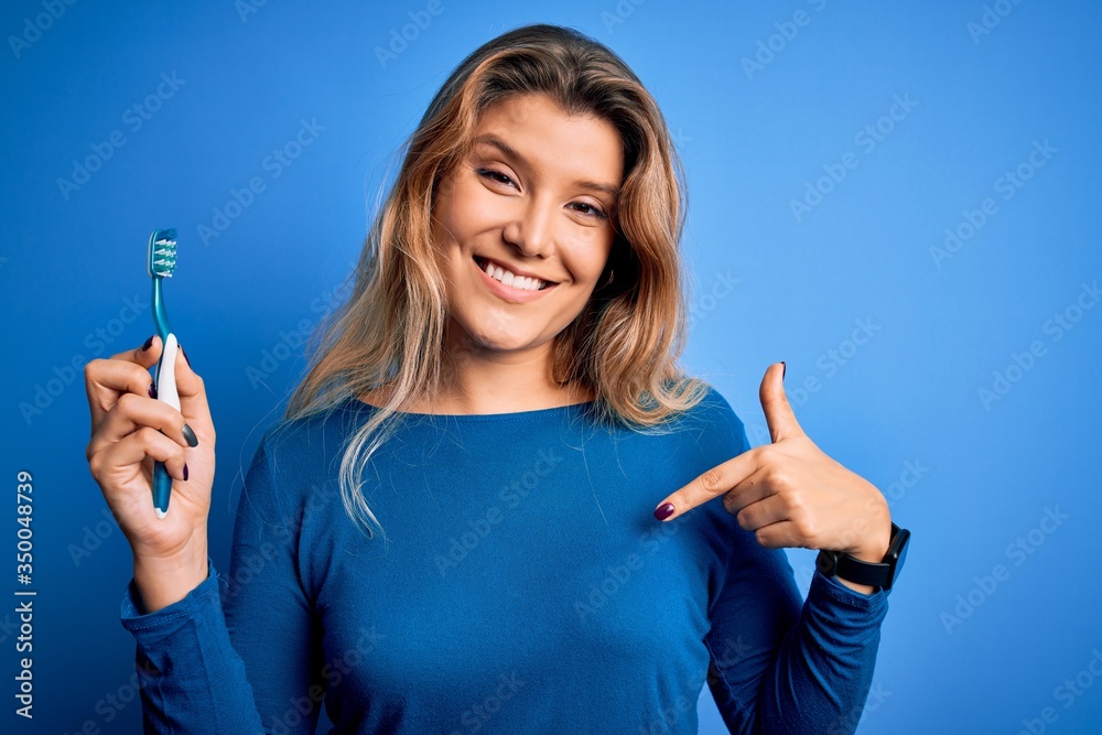 Young beautiful blonde woman using teeth brush over isolated blue background with surprise face pointing finger to himself Young beautiful blonde woman using teeth brush over isolated blue background with surprise face pointing finger to himself