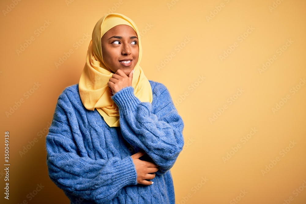 Young African American afro woman wearing muslim hijab over isolated yellow background with hand on chin thinking about question, pensive expression. Smiling with thoughtful face. Doubt concept. Young African American afro woman wearing muslim hijab over isolated yellow background with hand on chin thinking about question, pensive expression. Smiling with thoughtful face. Doubt concept.