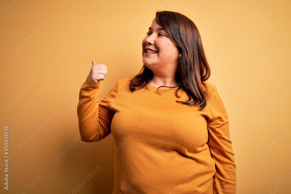 Beautiful brunette plus size woman wearing casual sweater over isolated yellow background smiling with happy face looking and pointing to the side with thumb up. Beautiful brunette plus size woman wearing casual sweater over isolated yellow background smiling with happy face looking and pointing to the side with thumb up.