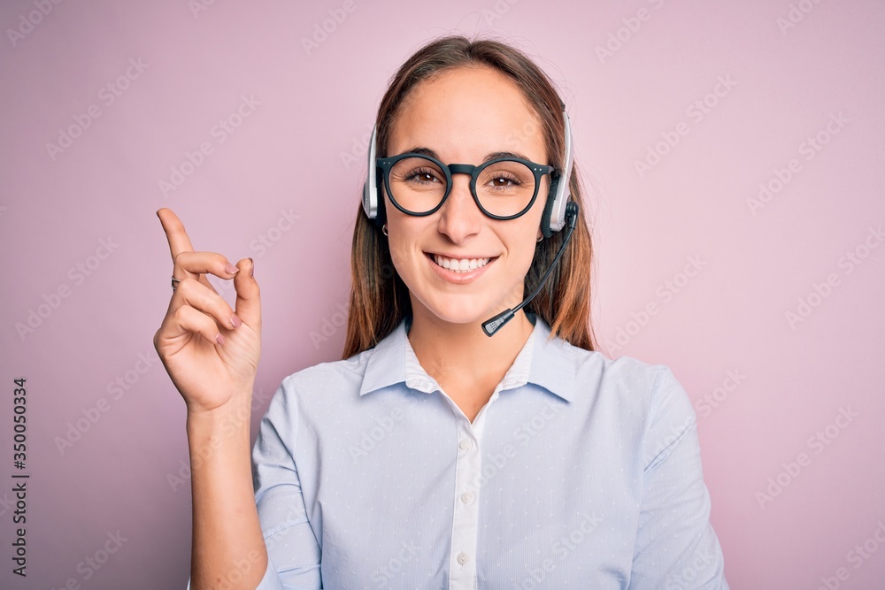 Young beautiful call center agent woman wearing glasses working using headset with a big smile on face, pointing with hand and finger to the side looking at the camera. Young beautiful call center agent woman wearing glasses working using headset with a big smile on face, pointing with hand and finger to the side looking at the camera.