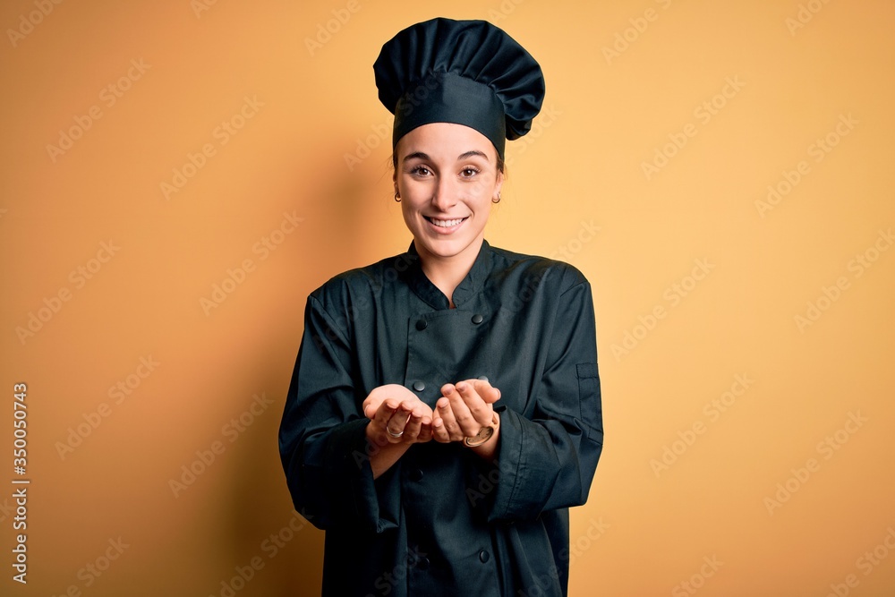 Young beautiful chef woman wearing cooker uniform and hat standing over yellow background Smiling with hands palms together receiving or giving gesture. Hold and protection