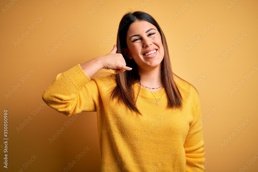 Young beautiful woman wearing casual sweater over yellow isolated background smiling doing phone gesture with hand and fingers like talking on the telephone. Communicating concepts. Young beautiful woman wearing casual sweater over yellow isolated background smiling doing phone gesture with hand and fingers like talking on the telephone. Communicating concepts.
