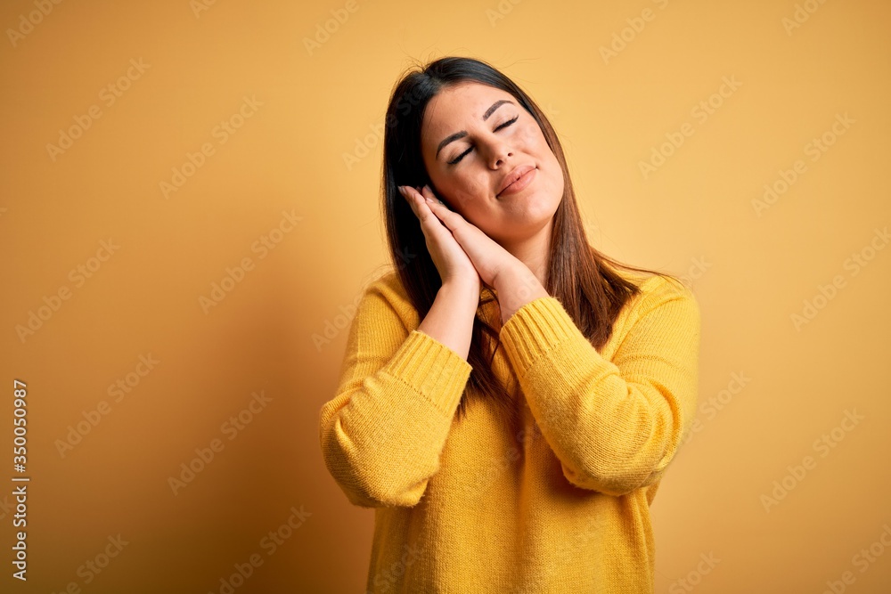 Young beautiful woman wearing casual sweater over yellow isolated background sleeping tired dreaming and posing with hands together while smiling with closed eyes. Young beautiful woman wearing casual sweater over yellow isolated background sleeping tired dreaming and posing with hands together while smiling with closed eyes.
