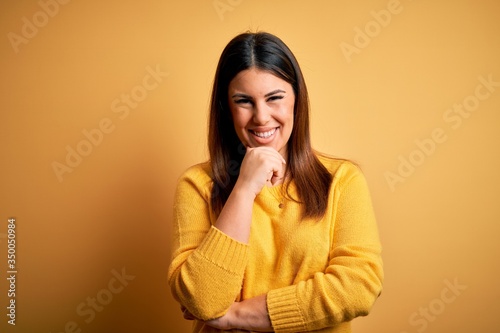 Young beautiful woman wearing casual sweater over yellow isolated background looking confident at the camera smiling with crossed arms and hand raised on chin. Thinking positive.