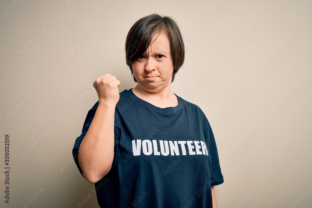 Young down syndrome volunteer woman wearing social care charity t-shirt angry and mad raising fist frustrated and furious while shouting with anger. Rage and aggressive concept. Young down syndrome volunteer woman wearing social care charity t-shirt angry and mad raising fist frustrated and furious while shouting with anger. Rage and aggressive concept.
