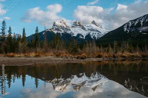 Breathtaking view of the iconic Three Sisters mountain peaks reflection in Policeman's Creek calm water while sunset. Canmore, Alberta, Canada. © Dajahof