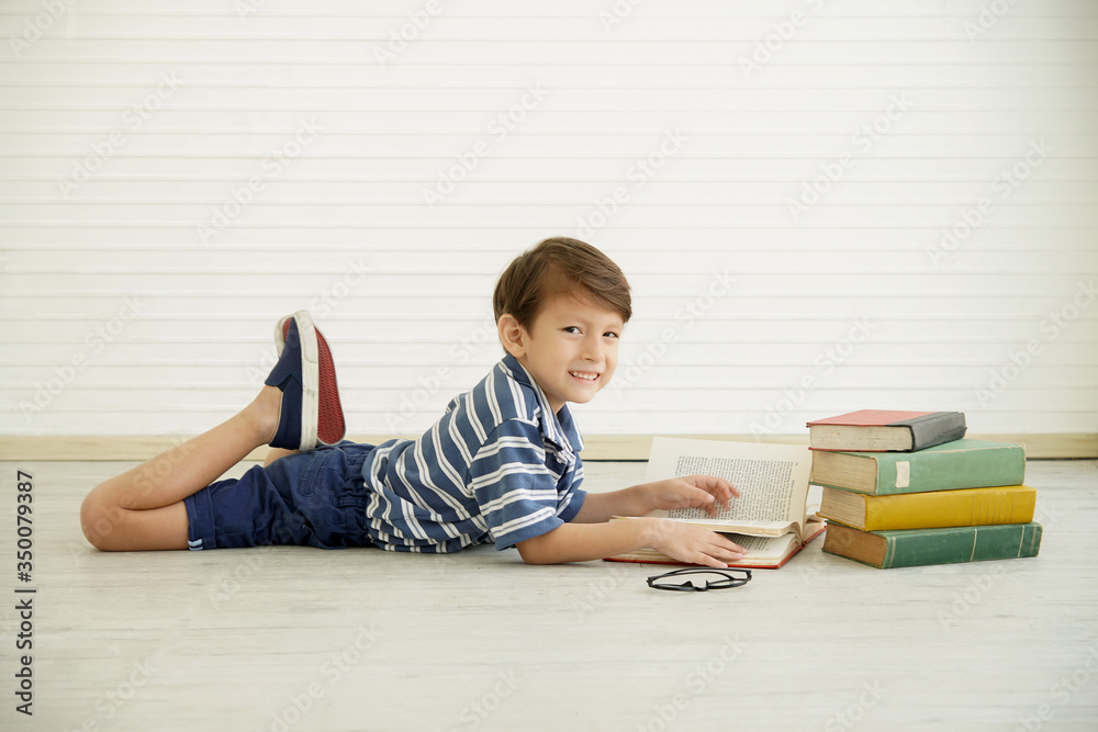 Portrait of little happy Caucasian boy lying raise both legs up on the floor looking at camera and reading a big book at home. There are many books and glasses beside him Portrait of little happy Caucasian boy lying raise both legs up on the floor looking at camera and reading a big book at home. There are many books and glasses beside him