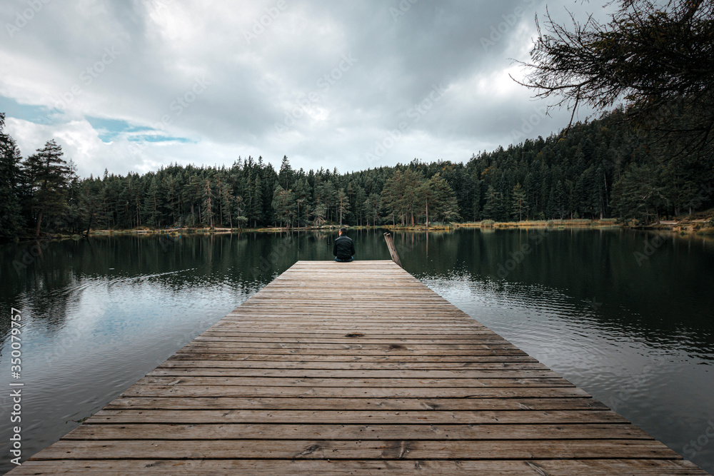  man sitting on the end of a pier jetty looking out to the water in a lake in a forest on a gloomy cloudy day