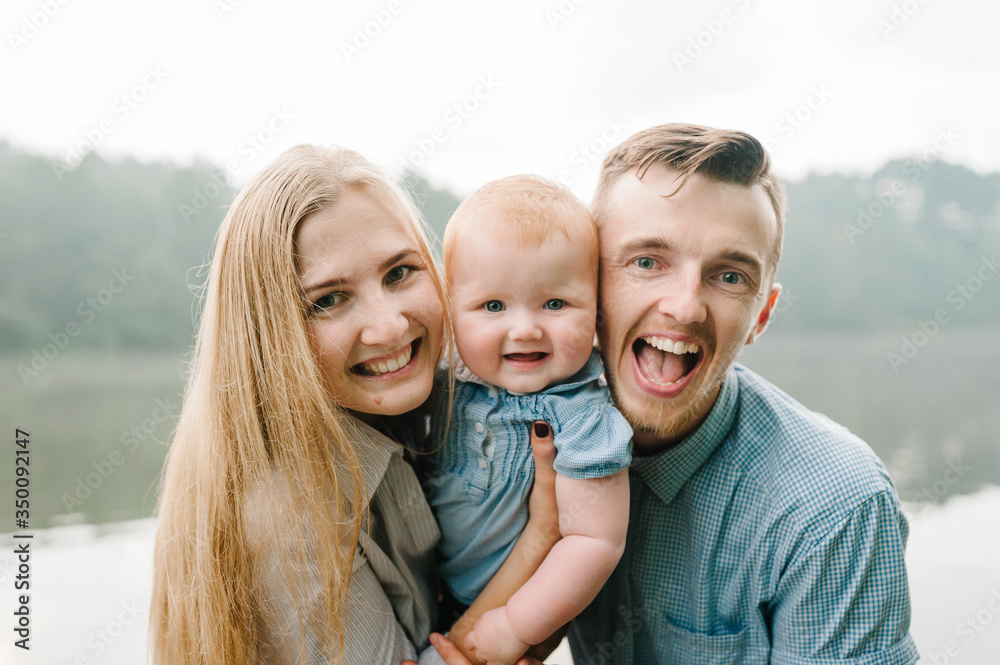 Fototapeta premium Happy young family near lake, pond. Family enjoying life together at meadow. People having fun in nature. Family bonds outside. Mother, father, child of three smiling while spending free time outdoors