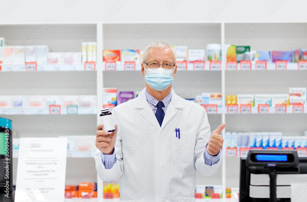 medicine, healthcare and people concept - senior apothecary wearing face protective medical mask for protection from virus disease with drug at pharmacy cash register showing thumbs up
