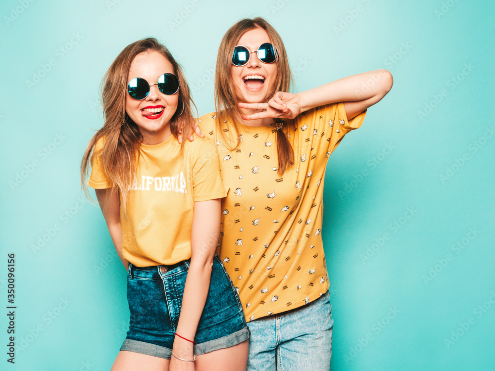 Two young beautiful smiling hipster girls in trendy summer yellow T-shirts. Sexy carefree women posing near blue wall. Trendy and positive models having fun in sunglasses