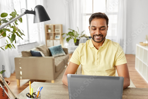 technology, remote job and lifestyle concept - happy indian man with laptop computer working at home office